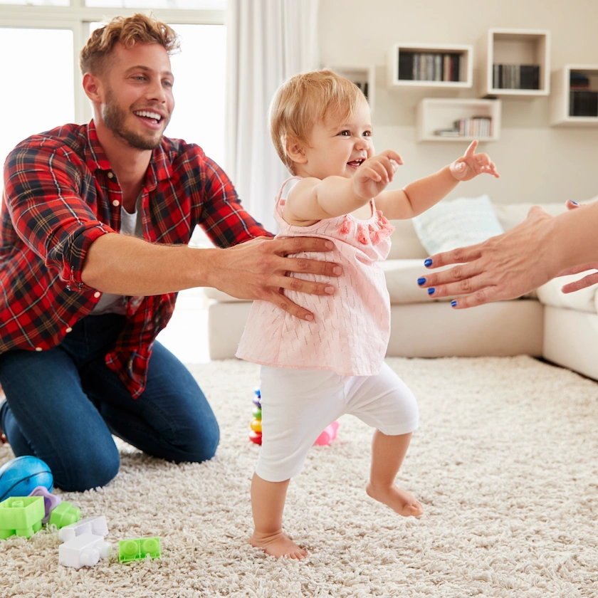 A toddler taking steps across a rug, arms out, while a kneeling man steadies her at the waist and another adult reaches toward her.